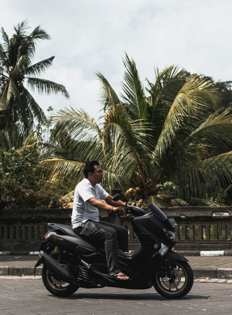 Man riding a black scooter NMAX (yamaha) through Kuta, Lombok, surrounded by tropical palm trees and traditional stone walls.